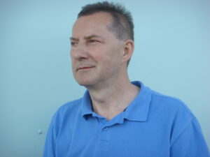 Portret mężczyzny patrzącego w lewą stronę. Artysta jest ubrany w niebieską koszulkę, jest mężczyzną w średnim wieku, ma krótkie, ciemne włosy.