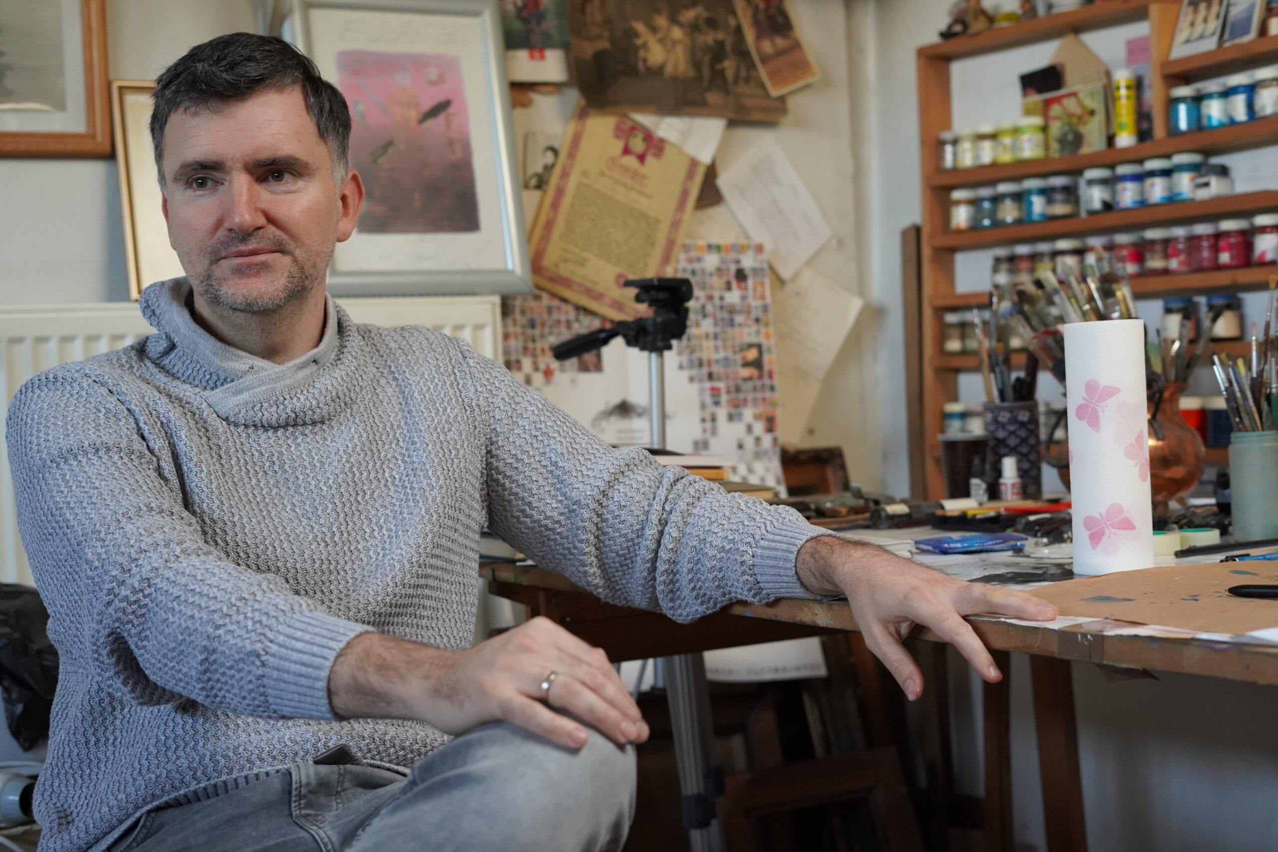 Portret artysty Piotra Nowińskiego w swojej pracowni. Siedzi przy stole, za nim obrazy i półka z farbami. Mężczyzna opiera się jedną  ręką o stół, druga leży na kolanie, które jest założone na drugie. Ma krótko ścięte ciemne włosy, z lekką siwizną. 