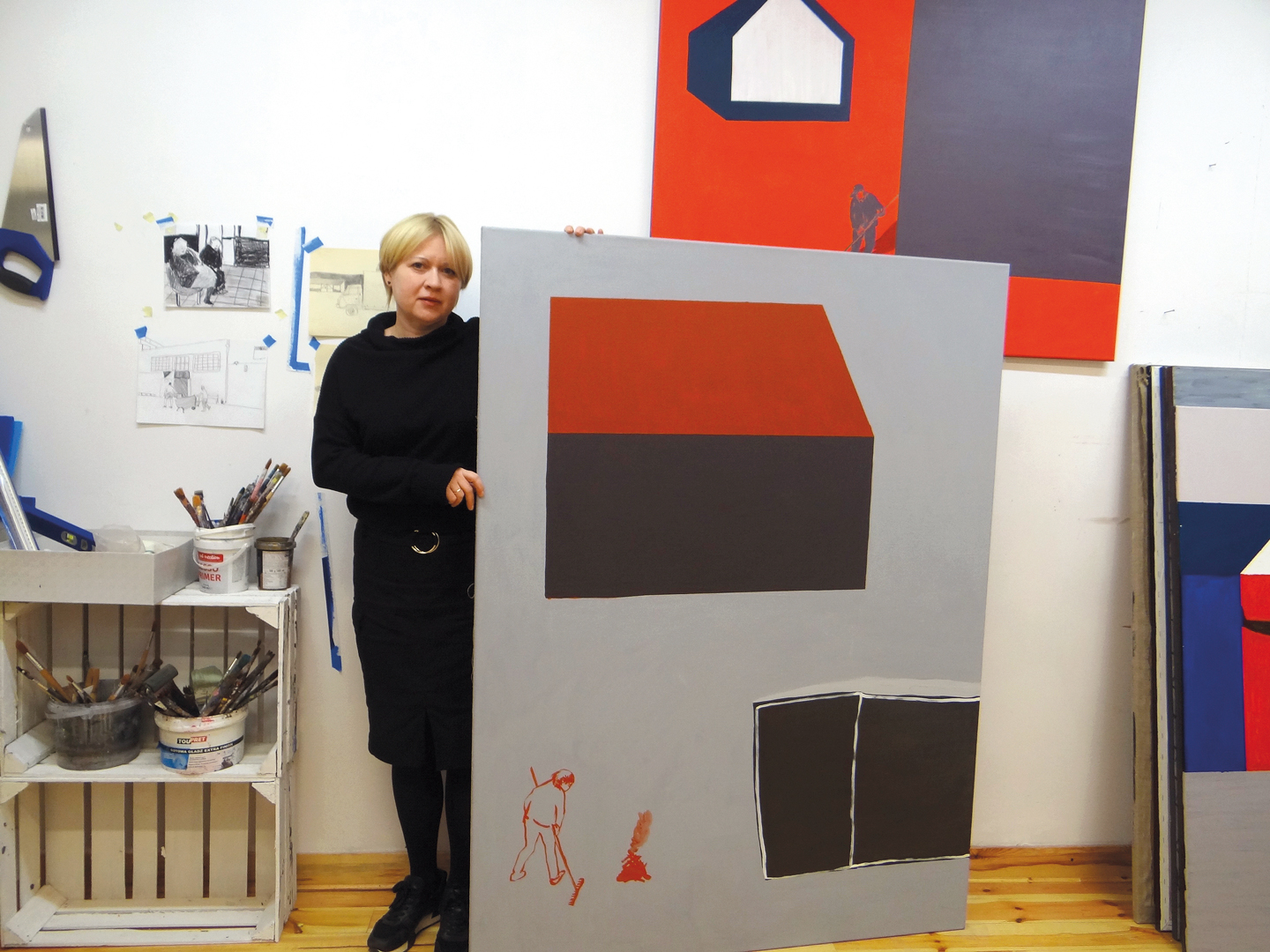 Artystka Joanna Mrozowska w swojej pracowni.  Ubrana jest na czarno, blondynka, stoi obok obrazu, który jest jej wzrostu. Na obrazie symbolicznie malowany domek z czerwonym dachem. Po lewej stronie szkicowo malowana postać grabiąca liście.