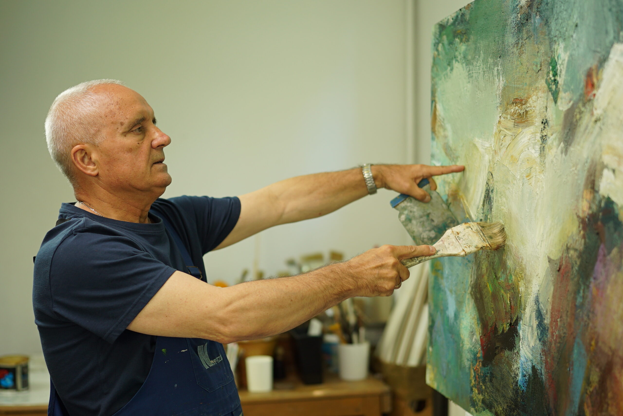 Na zdjęciu Artysta Ryszard Pasikowski, Który tworzy właśnie swój obraz. Artysta ubrany w granatową koszulkę i fartuch widoczny od pasa w górę. Ma siwe krótkie włosy. Po prawej stronie jest malowany abstrakcyjny obraz w kolorach zieleni i błękitu.