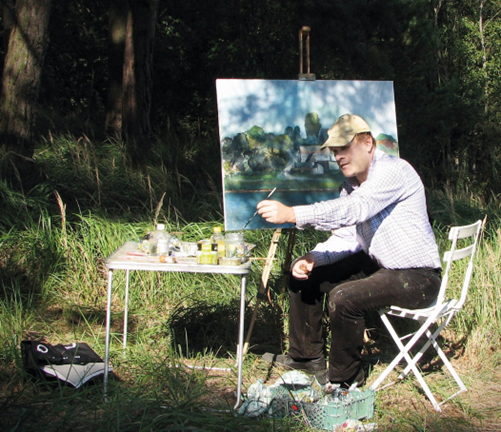 Artysta Maciej Mazurek Malujący w plenerze. Artysta Ma na sobie jasną koszulę, czapke z daszkiem i ciemne spodnie ubrudzone farbą. Siedzi przy sztaludze i miesza farby na stoliku.