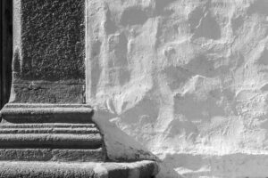 Fotografia czarno-biała, po lewej schody, po prawej biała, nieregularna, kamienna ściana.