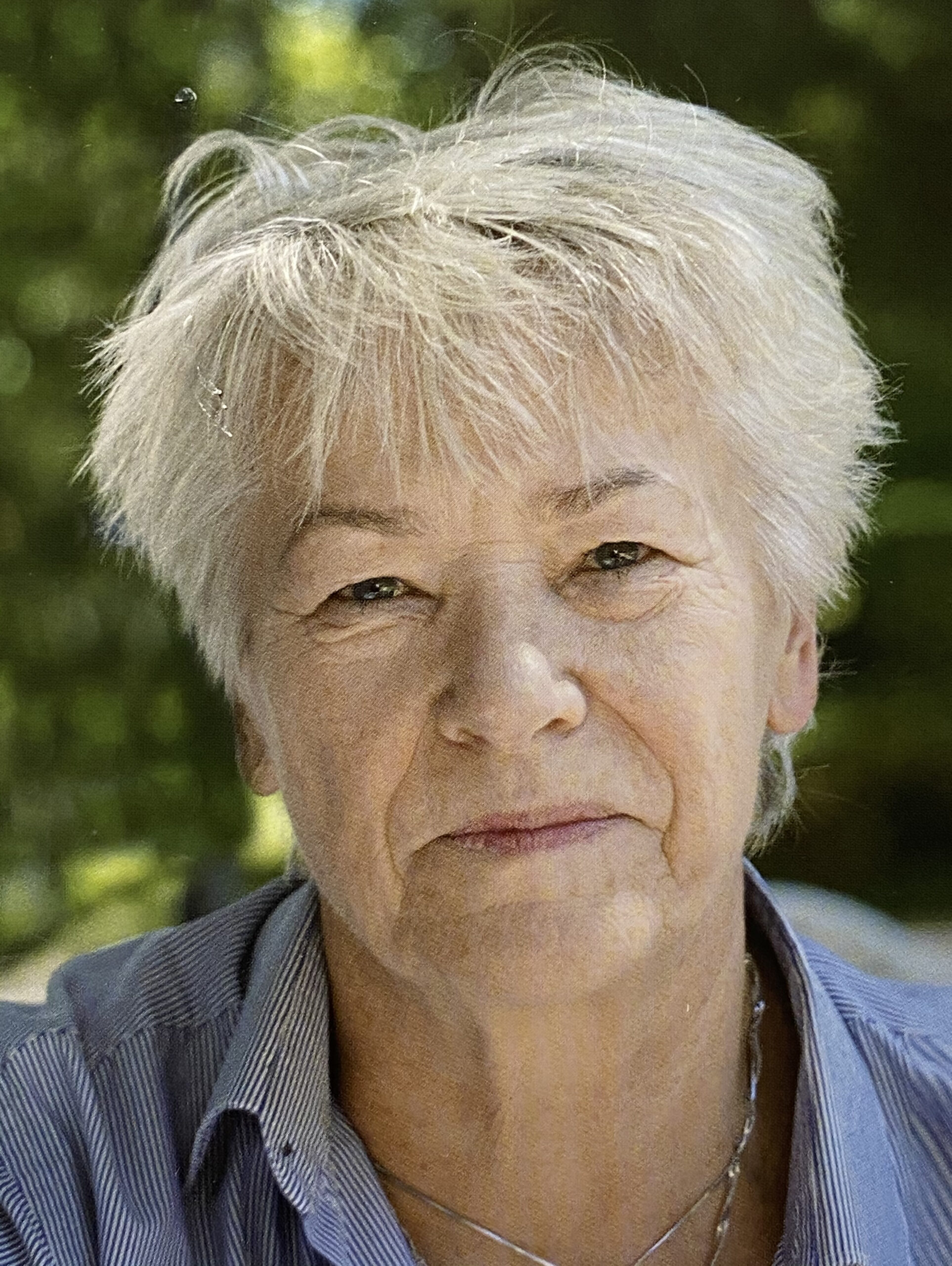 Portret artystki, starsza kobieta, krótko ścięta z jasnymi włosami patrzy na odbiorcę, ubrana w niebieską koszulę. W tle zieleń natury.