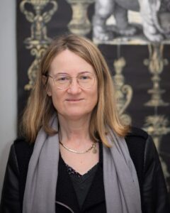 na Zdjęciu portret AGNIESZKA CIESLINSKA - KAWECKA. Dojrzała kobieta o blond włosach do ramion, w okularach. Ubrana w czarną bluzkę, na ramionach ma szary szalik