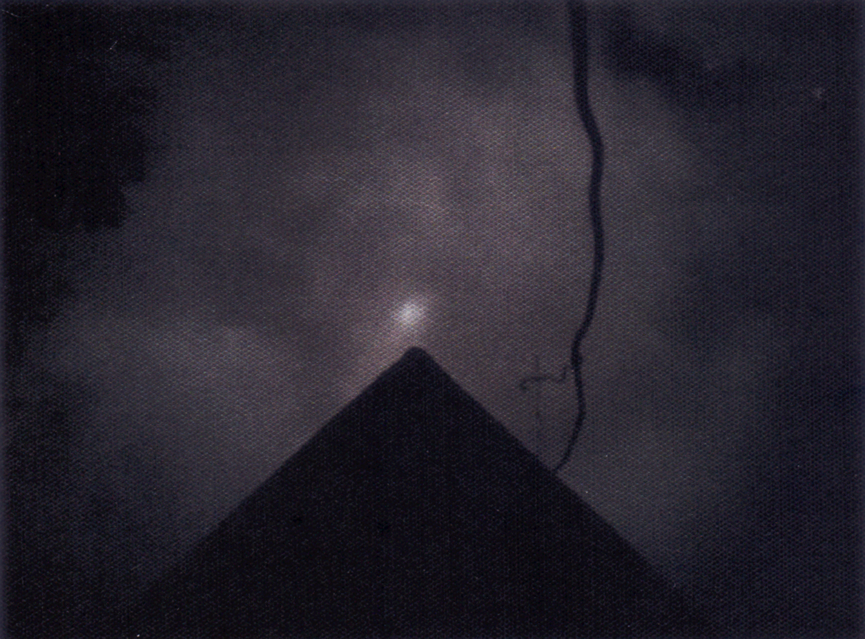 Na fotografii widać  piramidę. Nad nią jest jasny, zamglony punkt. Po prawej stronie zwisa kształt przypominający gałąź. Na pora nocy, prawie ciemno. 