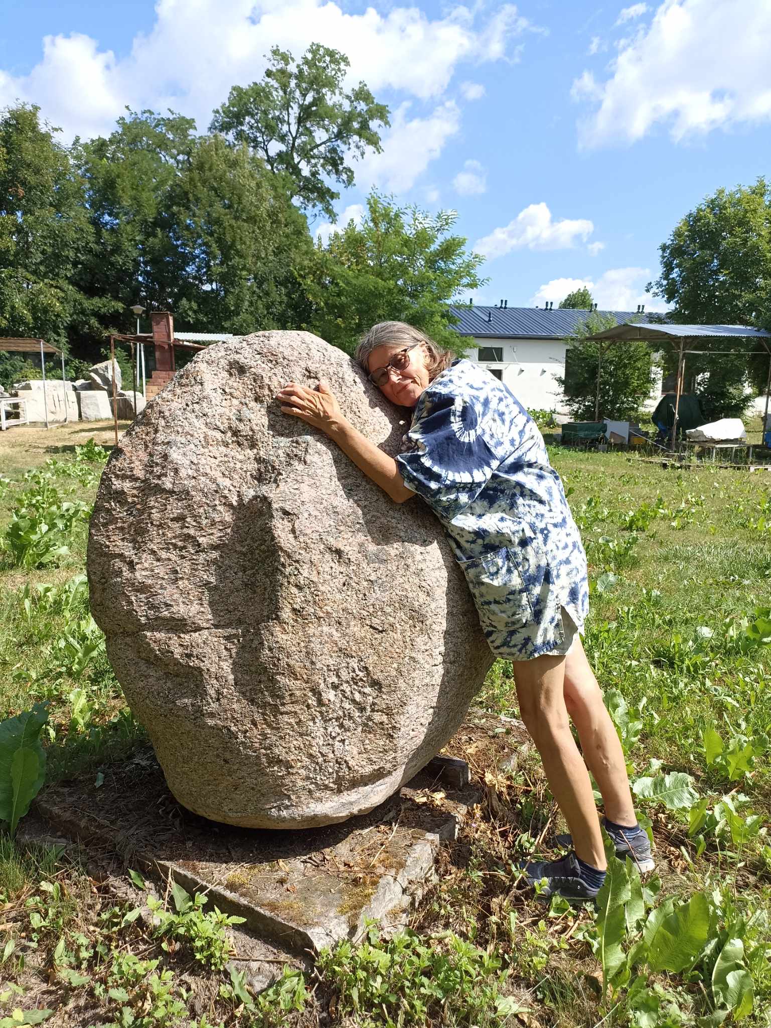 Artystka Jolanta Wołocznik opiera się, przytula do niego o kamień prawie jej wysokości. W tle zabudowania i drzewa.