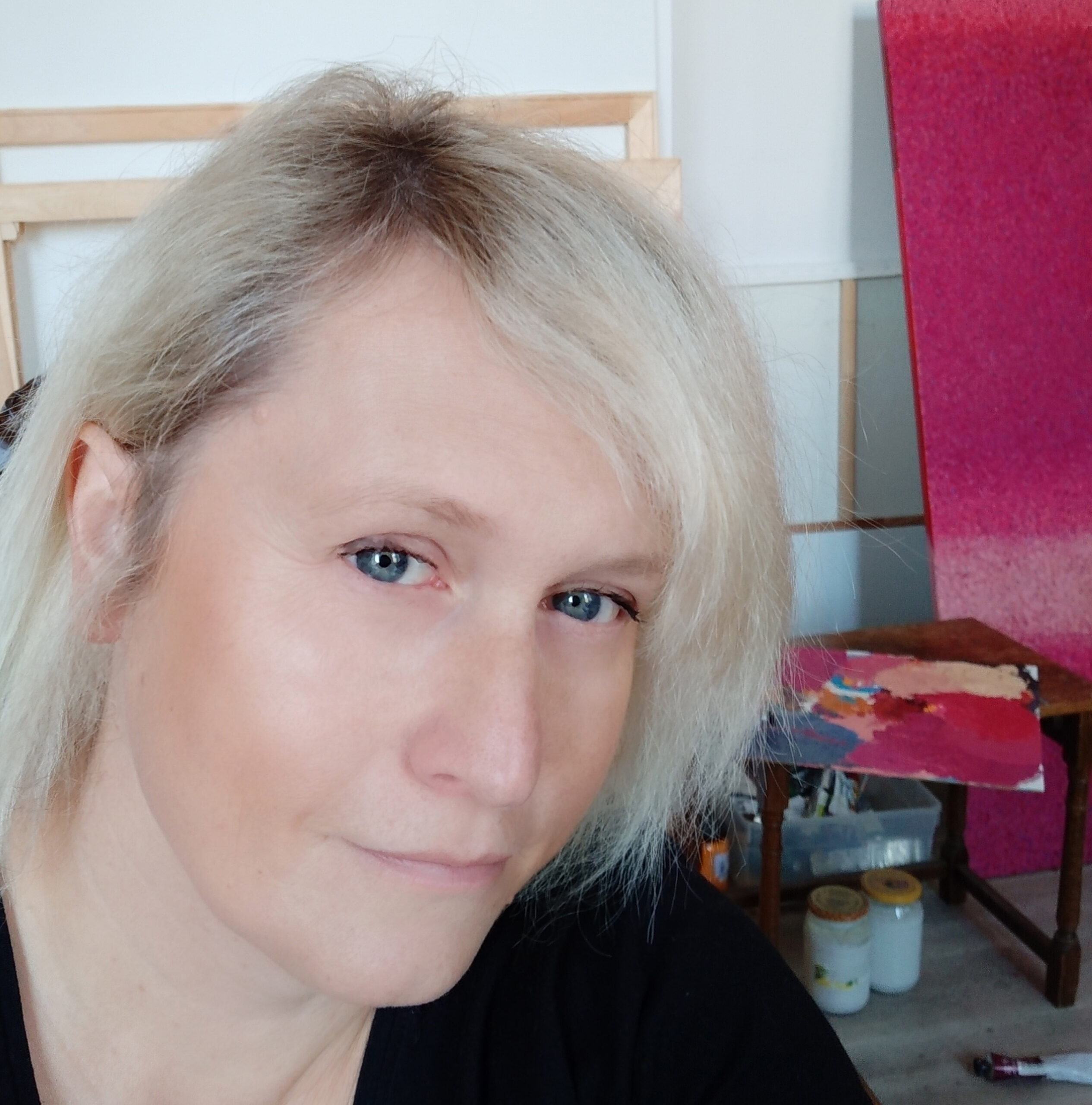 Na zdjęciu artystka Anna Wolska. Po lewej stronie widać ujęcie portretowe twarzy artystki. Anna Wolska patrzy wprost w kamerę i jest delikatnie uśmiechnięta. Ma wąskie, zaciśnięte usta. Artystka ma bardzo jasne włosy z przedziałkiem po prawej stronie. Głowa artystki jest lekko przechylona. W tle widać paletę z farbami, słoiki z zawartością w kolorze mlecznym. Paleta leży na ciemnobrązowym stołeczku o prostokątnym kształcie. Z tyłu głowy artystki widać oparte o białą ścianę blejtramy.